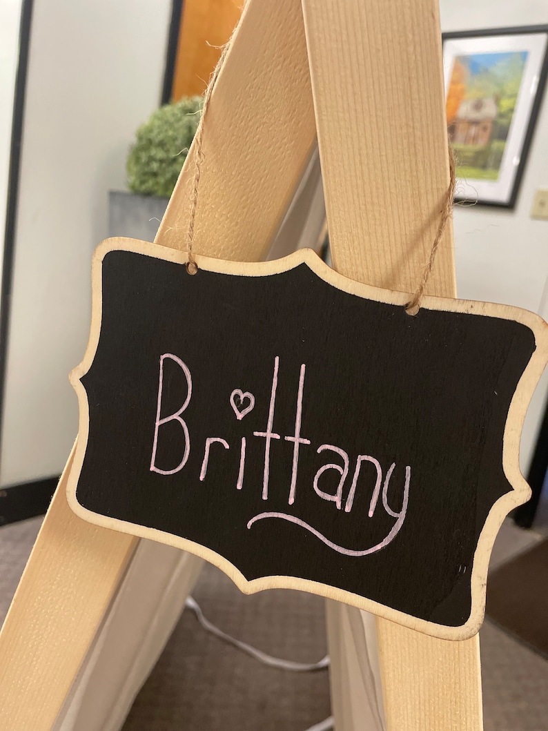May include: A black chalkboard sign with a wooden frame hangs from a string. The sign says "Brittany" in pink cursive with a small pink heart.
