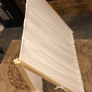 May include: A white fabric play tent with a wooden frame. The tent is set up on a patterned rug. The tent has white ribbons tied at the bottom of the fabric.