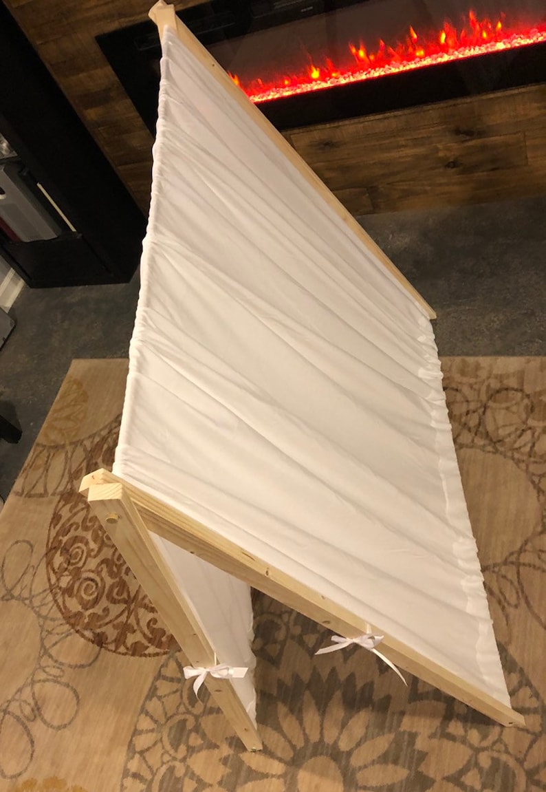 May include: A white fabric play tent with a wooden frame. The tent is set up on a patterned rug. The tent has white ribbons tied at the bottom of the fabric.