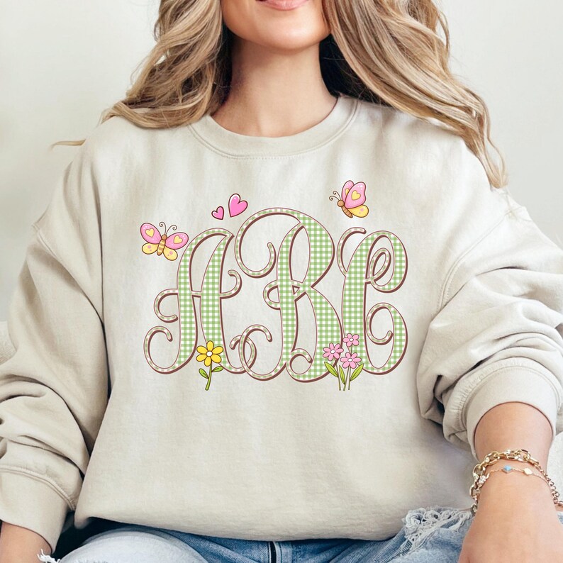 May include: A beige sweatshirt with a monogram design. The letters "ARB" are in a brown, curly font and filled with a green and white gingham pattern. The design includes butterflies, flowers, and hearts.