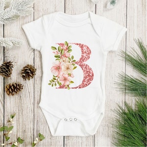Pu&ograve; includere: Body bianco per neonato con una lettera B glitter rosa e un design floreale rosa.