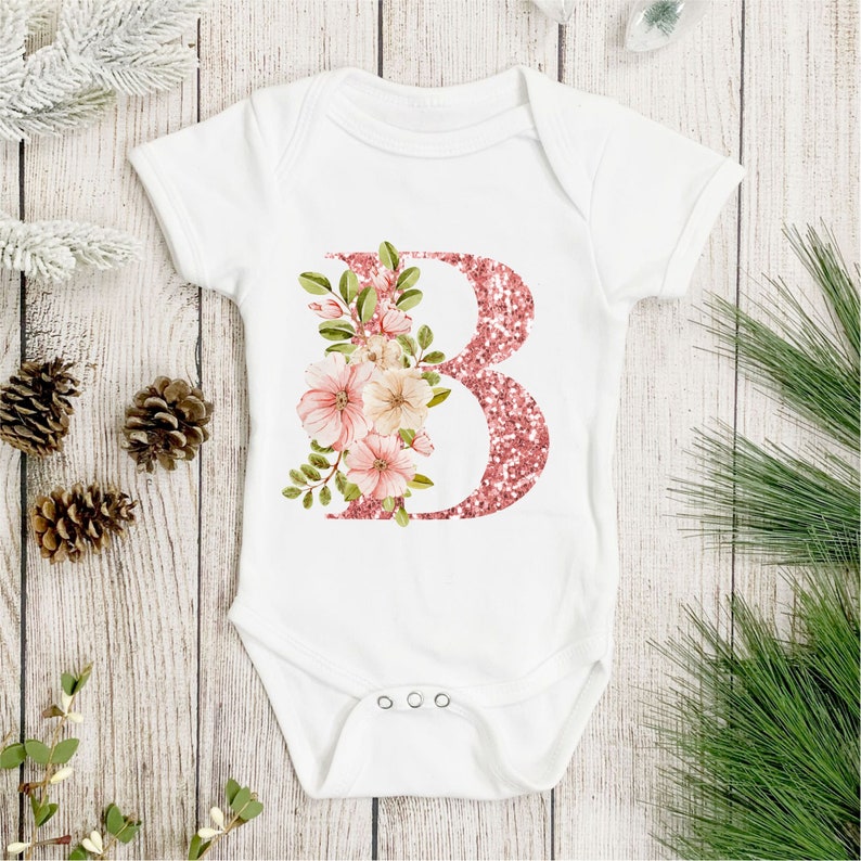 Pu&ograve; includere: Body bianco per neonato con una lettera B glitter rosa e un design floreale rosa.