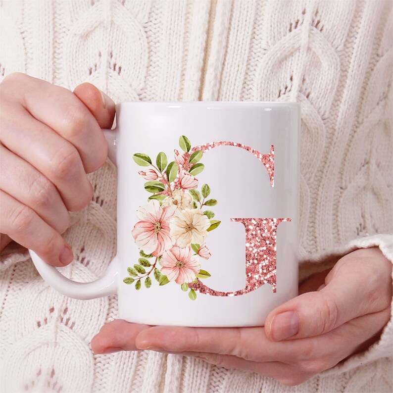 Pu&ograve; includere: Tazza in ceramica bianca con una lettera G di glitter rosa e un design floreale. I fiori sono rosa, bianchi e verdi.