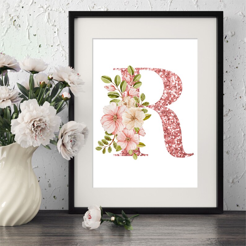 Pu&ograve; includere: Una stampa incorniciata con una lettera R di glitter rosa decorata con fiori rosa e bianchi. Lo sfondo &egrave; bianco.