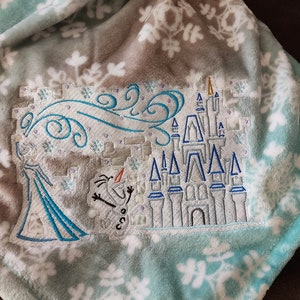 May include: A soft blue and grey fleece blanket with a white snowflake pattern. The blanket features an embroidered design of a castle, a snowman, and a swirling wind.