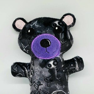 May include: A black plush teddy bear with a purple nose and black eyes. The bear is wearing a black and white fabric with a pattern of stars, moons, and spooky characters.