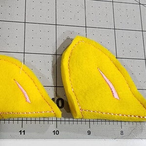 Puede incluir: Dos formas de orejas de tela de fieltro amarillo con costuras rosas. Las formas de las orejas están cosidas alrededor de los bordes y tienen una línea rosa en el centro.