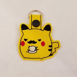 May include: Yellow Pikachu-themed keychain with black embroidered details. The keychain features a cartoon Pikachu with a mustache and a teacup. The keychain has a yellow strap and a silver keyring.