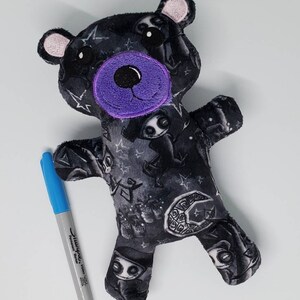 May include: A black plush teddy bear with a purple nose and eyes. The bear is covered in a black and white print of spooky characters and stars.