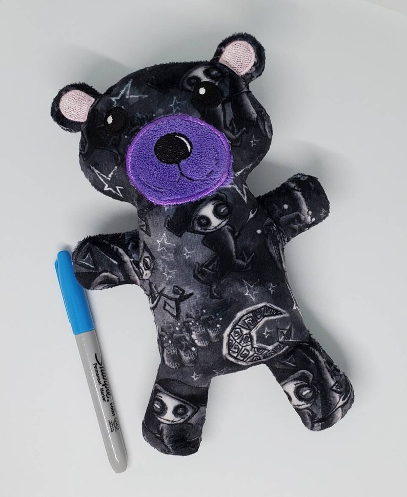 May include: A black plush teddy bear with a purple nose and eyes. The bear is covered in a black and white print of spooky characters and stars.
