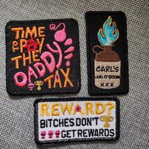 May include: Three embroidered patches with black borders. One square patch reads TIME TO PAY THE DADDY TAX in orange and pink with an anarchy symbol. One rectangular patch shows a jug labeled CARL'S JUG O BOOM XXX with blue flames. One white patch reads REWARD? BITCHES DON'T GET REWARDS.