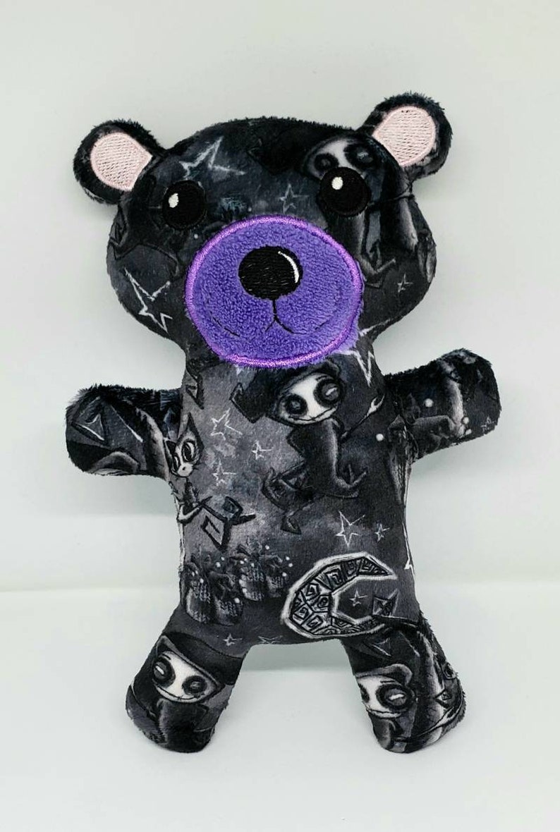 May include: A black plush teddy bear with a purple nose and black eyes. The bear is wearing a black and white fabric with a pattern of stars, moons, and spooky characters.