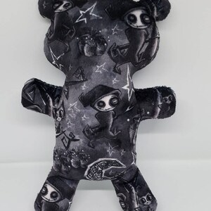 May include: A black plush teddy bear with a white and gray print of a spooky character with a hat and a crescent moon. The character is repeated throughout the fabric.