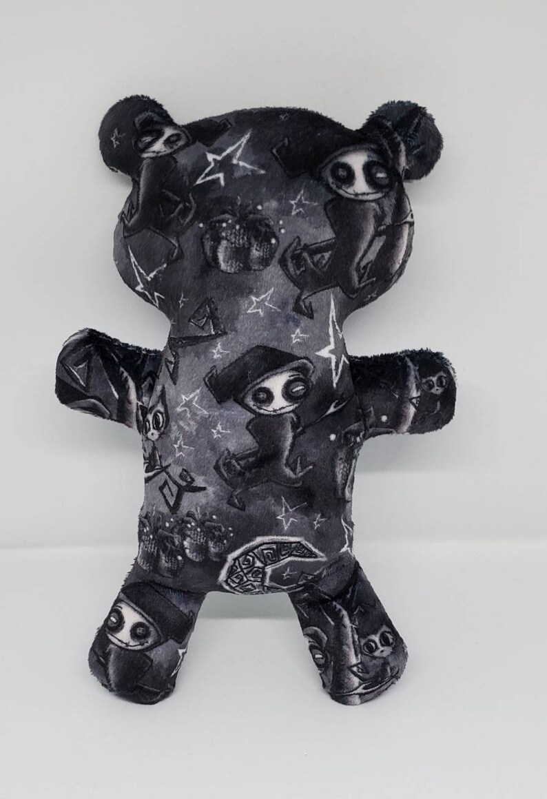 May include: A black plush teddy bear with a white and gray print of a spooky character with a hat and a crescent moon. The character is repeated throughout the fabric.
