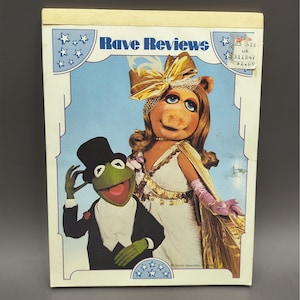 May include: A rectangular notepad titled Rave Reviews featuring a printed illustration of Miss Piggy in a white gown with a gold bow and Kermit the Frog in a black tuxedo and top hat. The border is light blue with white stars and a price sticker.