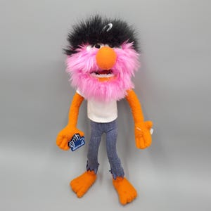May include: A pink and orange puppet with black hair and a white shirt. The puppet is wearing blue jeans with frayed hems. The puppet is holding a blue and white sticker that says "applause".