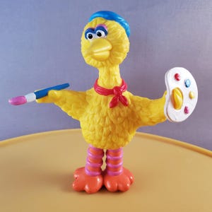 May include: A yellow Big Bird figurine wearing a blue beret and a red scarf, holding a paintbrush and a white paint palette with colorful paint dots.