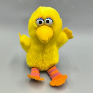 May include: A plush yellow bird toy with large blue and black eyes, a yellow beak, and orange feet. The bird has orange and pink striped legs and outstretched wings. The toy is a character from a children's television show.
