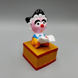 May include: A hand-painted figurine of a character with a pink face, black hair, and a white ruffled collar. The figure is seated on a yellow block with red trim and holds a white paper with red markings.