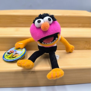 May include: A plush toy of the Muppet character Animal. The toy has a pink head, black hair, and orange nose. It wears a white shirt, black pants, and orange feet and hands. A small, round tag is next to the toy.