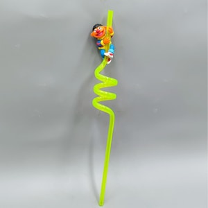 May include: A neon green, spiral-shaped drinking straw with a cartoon character figurine attached. The figurine is of a character with orange skin, brown hair, and colorful clothing. The straw is made of plastic and is designed for beverages.