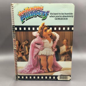 May include: A spiral-bound notebook featuring Miss Piggy on the cover with the text Jim Henson's MUPPETS and It's hard to be humble when you're absolutely GORGEOUS! The cover shows Miss Piggy in a white dress and pink ruffled shawl.