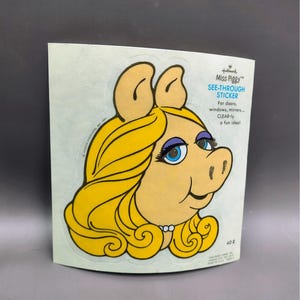 May include: A vintage Miss Piggy see-through sticker. The sticker features a cartoon illustration of Miss Piggy's face with blonde hair, blue eyes, and a pearl necklace. Text on the sticker reads "SEE-THROUGH STICKER" and "CLEAR-ly a fun idea!"