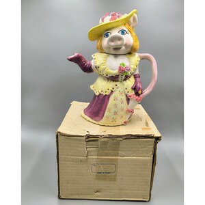 May include: A ceramic teapot shaped like a pig wearing a yellow hat and dress, with purple accents. The teapot is standing on a cardboard box. The teapot has a pink handle and a spout. The pig has blue eyes and is holding a small bouquet of flowers.