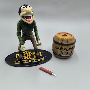 May include: A collectible action figure of a Muppet character, wearing a green jacket and brown pants, standing on a black base. A wooden barrel labeled "GUNPOWDER" and a red and white object are also present. The base reads "The Muppet Show 25 Years."