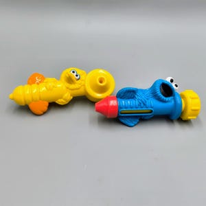 May include: Two colorful Sesame Street character-themed water guns. One is yellow with orange accents, resembling Big Bird. The other is blue with white eyes, resembling Cookie Monster. Both have red and yellow nozzles.