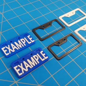 Puede incluir: Cuatro marcos para placas de matr&iacute;cula, dos negros y dos blancos, con dos placas de matr&iacute;cula azules que dicen "EXAMPLE" en letras blancas.