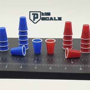 May include: Miniature blue and red plastic cups with white rims are arranged on a black surface. Some cups are stacked, while others are lying down. The image includes the text "1:12 SCALE".