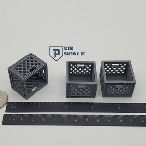 Puede incluir: Tres cajas de leche de pl&aacute;stico gris, cada una mide aproximadamente 5 cm de ancho, est&aacute;n dispuestas sobre una superficie blanca. Una regla se coloca debajo de las cajas, con marcas en pulgadas y cent&iacute;metros.