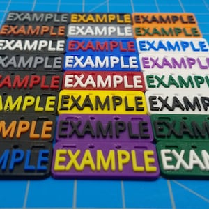 Puede incluir: Un conjunto de 10 etiquetas de pl&aacute;stico con la palabra "EXAMPLE" impresa en negro. Las etiquetas son de diferentes colores: gris, blanco, azul, morado, verde, amarillo, naranja, rojo y negro.