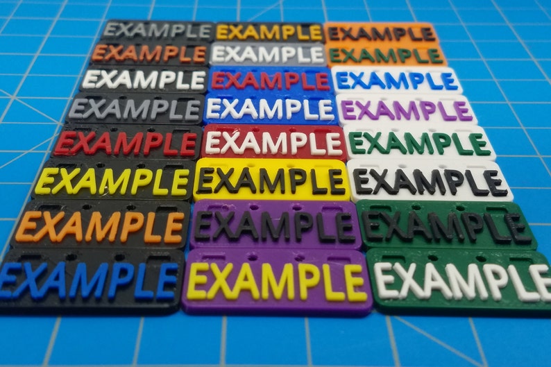 Puede incluir: Un conjunto de 10 etiquetas de pl&aacute;stico con la palabra "EXAMPLE" impresa en negro. Las etiquetas son de diferentes colores: gris, blanco, azul, morado, verde, amarillo, naranja, rojo y negro.