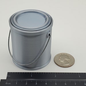 Pode incluir: Um pequeno pote de tinta de plástico cinza com uma alça de metal. O pote tem aproximadamente 5 cm de altura e está ao lado de uma moeda de 25 centavos de dólar americano para escala.