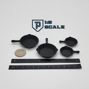 May include: Four miniature black cast iron skillets of varying sizes are displayed. A quarter and two rulers are next to the skillets. The text "1:6 SCALE" is visible in the upper right corner.