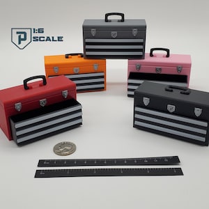 May include: Five 1:6 scale toolboxes in different colours: red, orange, grey, pink, and black. Each toolbox has three drawers and a handle on the top. The toolboxes are arranged in a semi-circle on a white surface. A ruler is in the bottom right corner of the image.