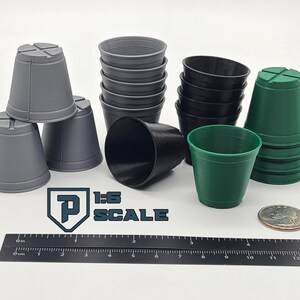 May include: A collection of small, plastic plant pots in various colors: gray, black, and green. The pots are arranged on a white surface with a ruler, a coin, and the text "1:6 SCALE".