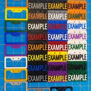 Puede incluir: Una colecci&oacute;n de 14 marcos de matr&iacute;cula de pl&aacute;stico en varios colores, incluyendo rojo, naranja, amarillo, verde, azul, morado, blanco y negro. Cada marco tiene dos agujeros para tornillos y la palabra "EXAMPLE" impresa en &eacute;l.