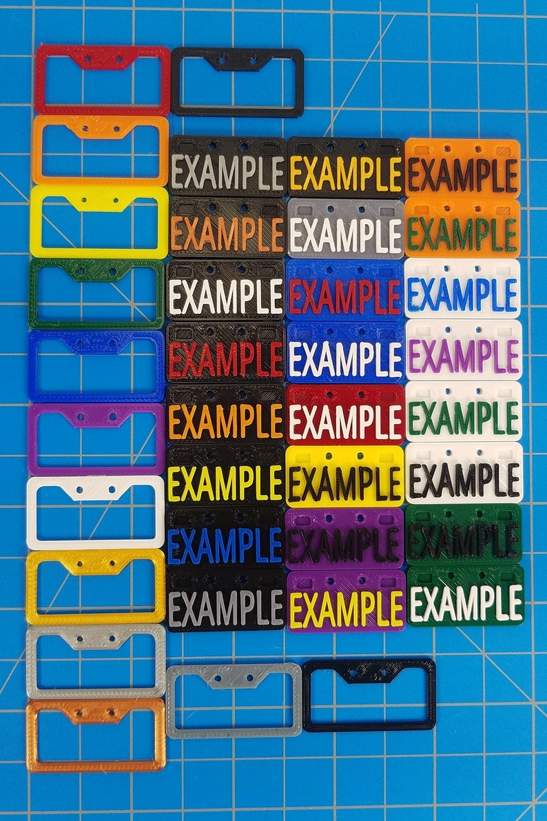 Puede incluir: Una colecci&oacute;n de 14 marcos de matr&iacute;cula de pl&aacute;stico en varios colores, incluyendo rojo, naranja, amarillo, verde, azul, morado, blanco y negro. Cada marco tiene dos agujeros para tornillos y la palabra "EXAMPLE" impresa en &eacute;l.