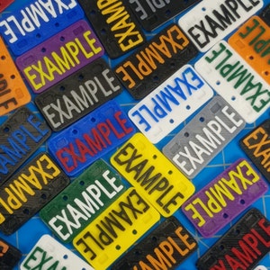 Puede incluir: Primer plano de una variedad de peque&ntilde;as placas de matr&iacute;cula de pl&aacute;stico con la palabra "EXAMPLE" impresa en ellas en diferentes colores y fuentes. Las placas est&aacute;n dispuestas en un patr&oacute;n aleatorio sobre una superficie azul.