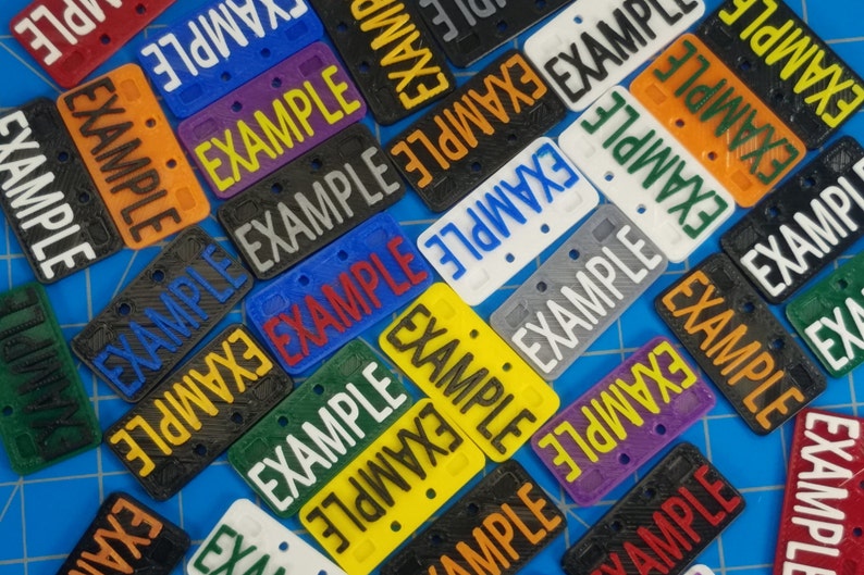 Puede incluir: Primer plano de una variedad de peque&ntilde;as placas de matr&iacute;cula de pl&aacute;stico con la palabra "EXAMPLE" impresa en ellas en diferentes colores y fuentes. Las placas est&aacute;n dispuestas en un patr&oacute;n aleatorio sobre una superficie azul.