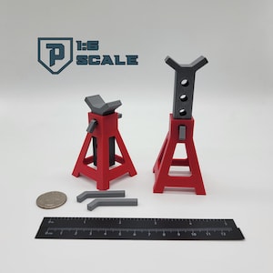 May include: Red and gray 1:6 scale model jack stands, with a ruler and a coin for size comparison. The jack stands have a triangular base and a height-adjustable top. The text "1:6 SCALE" is visible in the background.
