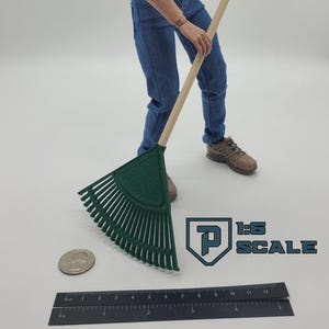 May include: A green plastic rake with a wooden handle. The rake is held by a person wearing blue jeans and brown shoes. A ruler and a quarter are on the ground. The text "1:6 SCALE" is visible.