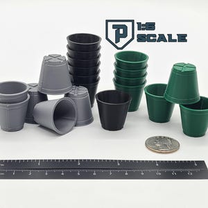 May include: A collection of small, plastic plant pots in various colors: gray, black, and green. A ruler with inch and centimeter markings is placed next to the pots. The text "1:6 SCALE" is visible in the upper right corner.