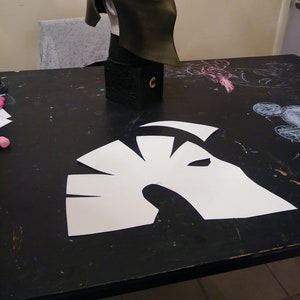 Op de afbeelding: Een zwarte en gouden helm met een wit papieren sjabloon van de helm op een zwarte tafel.