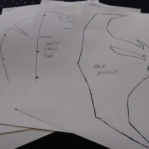 Puede incluir: Cuatro hojas de papel blanco con dibujos de tinta negra de un diseño de casco. El papel está etiquetado como "Spawn" y "Main Shell Part" y "Face Accent".