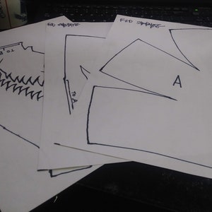 Puede incluir: Tres patrones de costura en papel blanco, etiquetados como "Delantero", "Pliegue" y "Forma de pliegue". Los patrones están dibujados con tinta negra e incluyen marcas para "A" y "B".