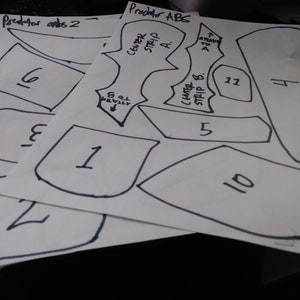 May include: A set of paper patterns for a Predator costume. The patterns are numbered 1 through 11 and include instructions for attaching the pieces. The text on the patterns reads "Predator ABS" and "Predator Predator ants 2".